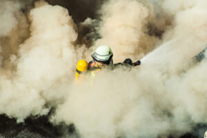 Feuerwehrman mit Wasserschlauch in einer gelben Rauchwolke